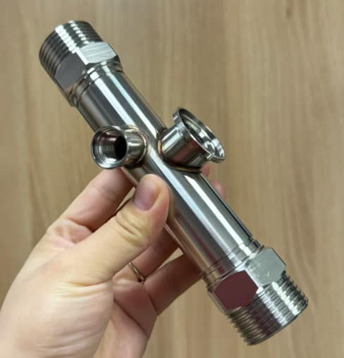 Stainless steel manifold held in hand showing CNC machined threads and TIG welded ports with visible heat-affected zones.