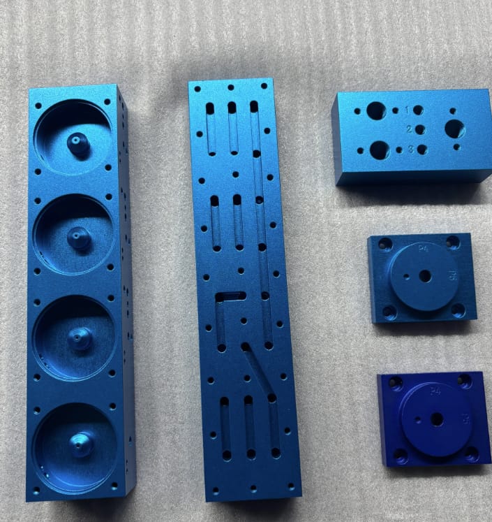 Custom blue anodized aluminum CNC machined parts including slotted plates, sensor housings, and mounting blocks with bead blasted finish.