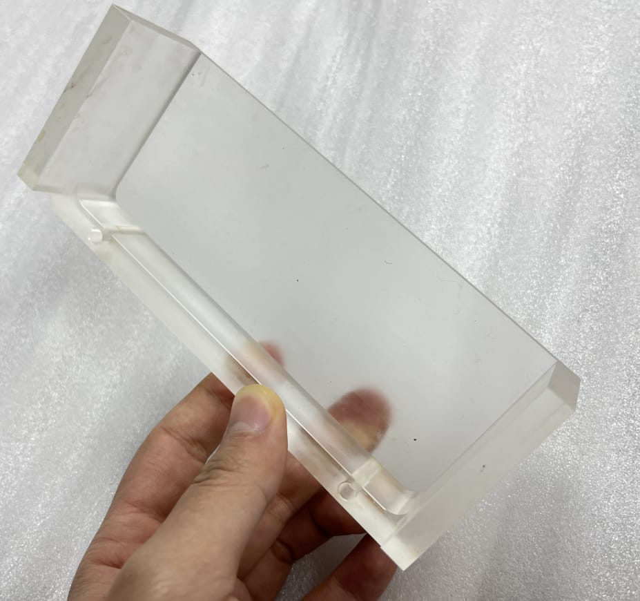 Clear CNC machined acrylic block held in hand showing internal milled pockets and drilled channels with matte finish before polishing.