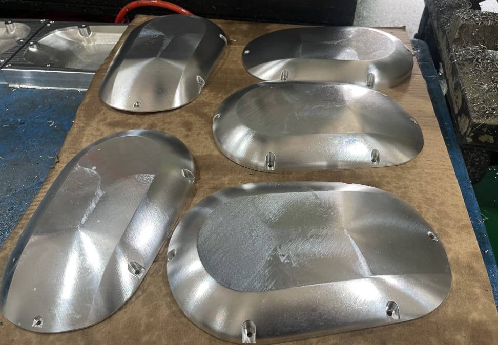Batch of five aluminum covers with complex organic curvature showing machined tool paths on a workbench at Rapid Model factory.